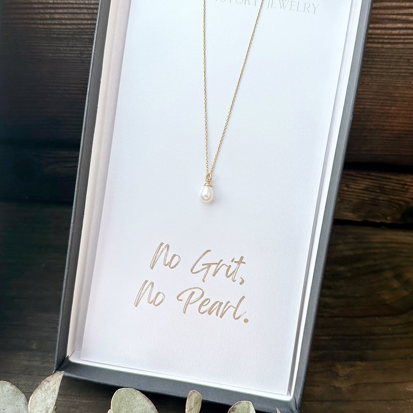 Gold necklace on a card with 'No Grit, No Pearl' text on a wooden surface