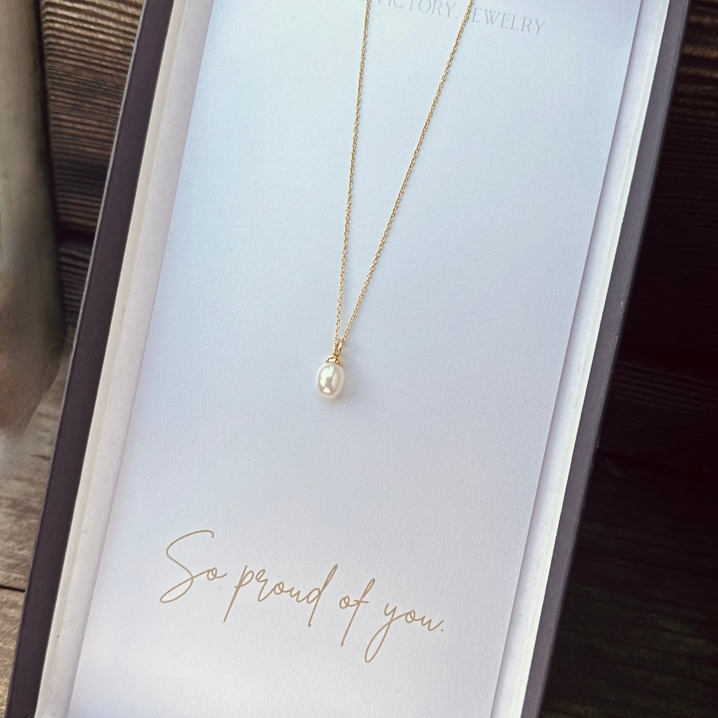 Gold necklace with a pearl pendant on a card that says 'So proud of you' on a wooden surface.