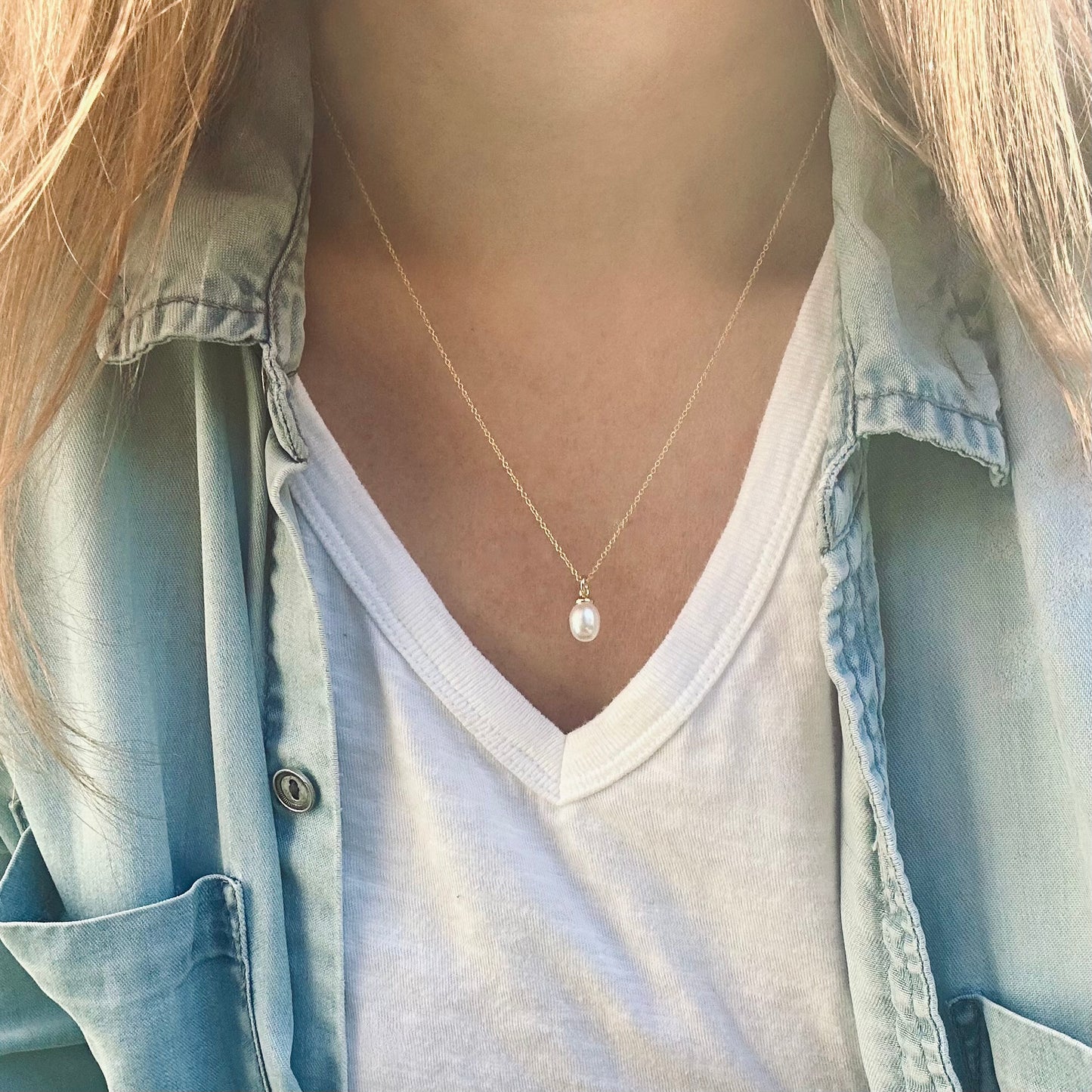 Person wearing a gold necklace with a pearl pendant, dressed in a light blue denim shirt.